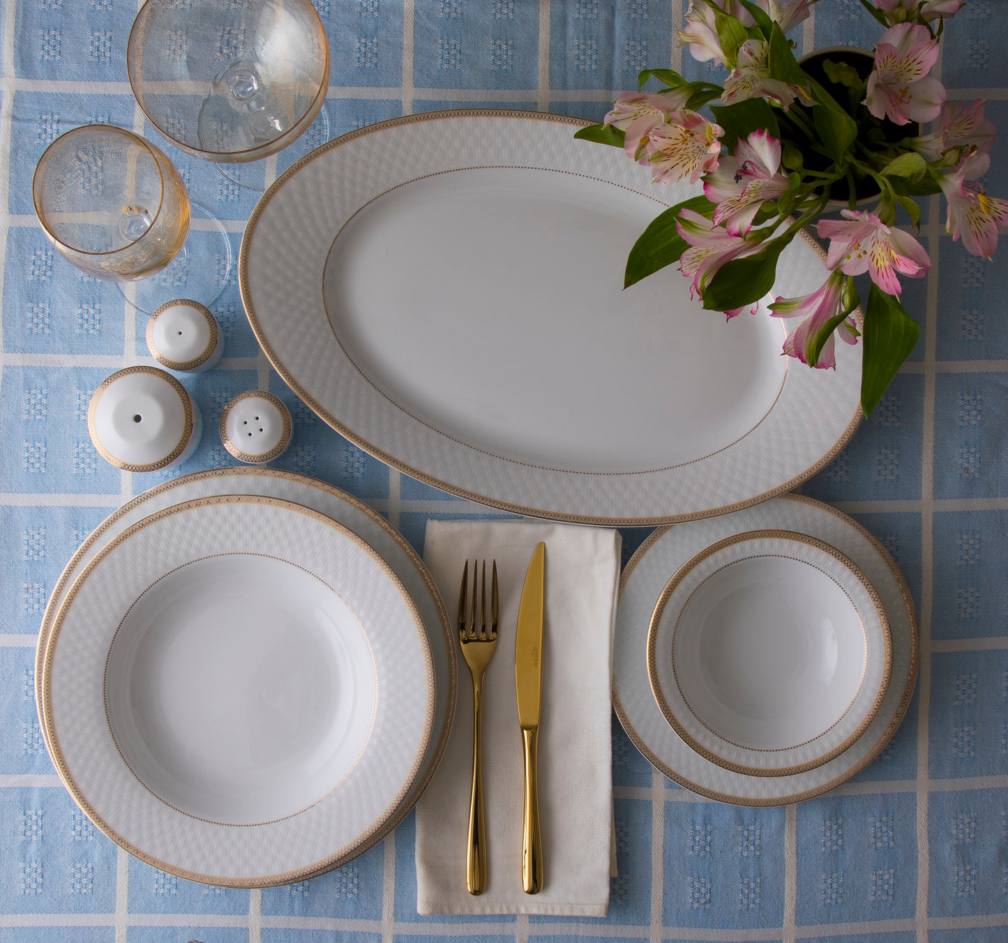 Sepinood-serviset dekket på en lyseblå og hvitrutete duk med ovalt serveringsfat, tallerkener, skåler, gullfarget bestikk, salt- og pepperbøsser og friske blomster